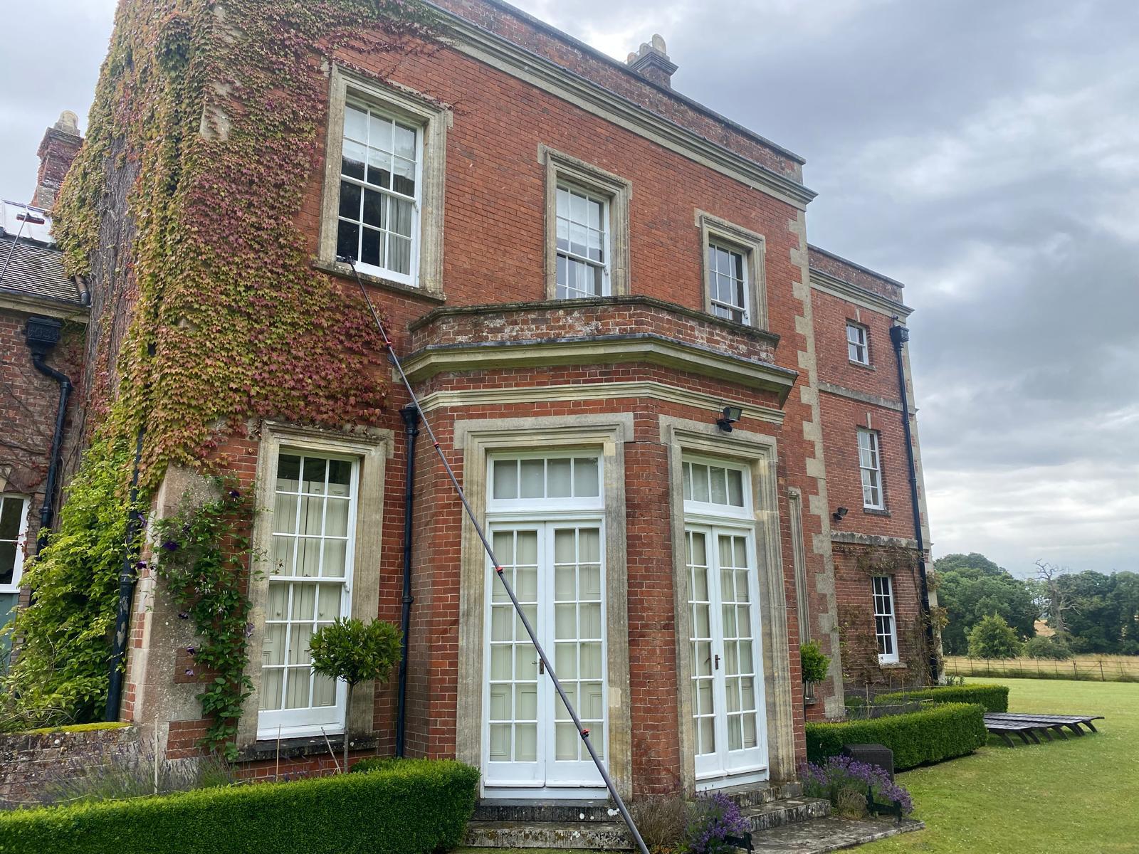 A large red-brick house with white-trimmed windows and doors, partially covered in ivy. A long pole hints at recent high level cleaning. Greenery and fields are visible in the background, adding to the home's charm.
