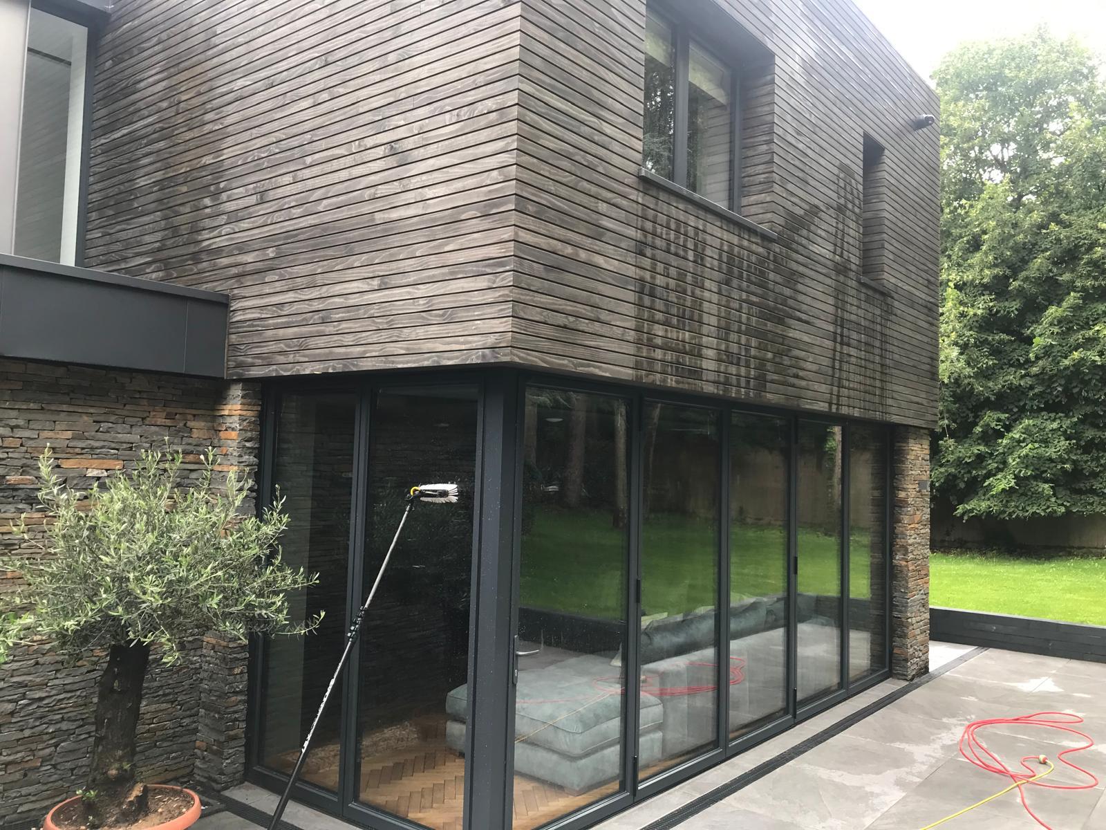 Modern house exterior with large floor-to-ceiling glass windows, dark wood cladding, and a stone wall. A spotless patio hints at expert driveway jet wash services, while a lush garden is visible through the freshly cleaned windows.