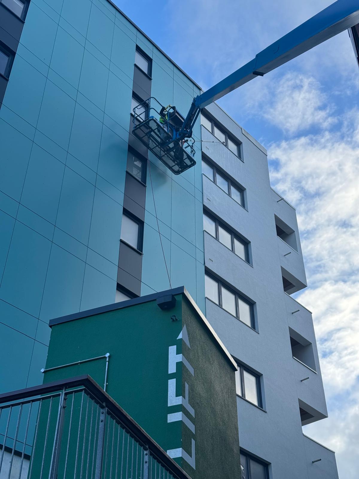 A worker in a lift is performing high level cleaning or inspecting the windows of a modern, multi-story building under a partly cloudy sky. The lift extends from the ground to the upper floors.