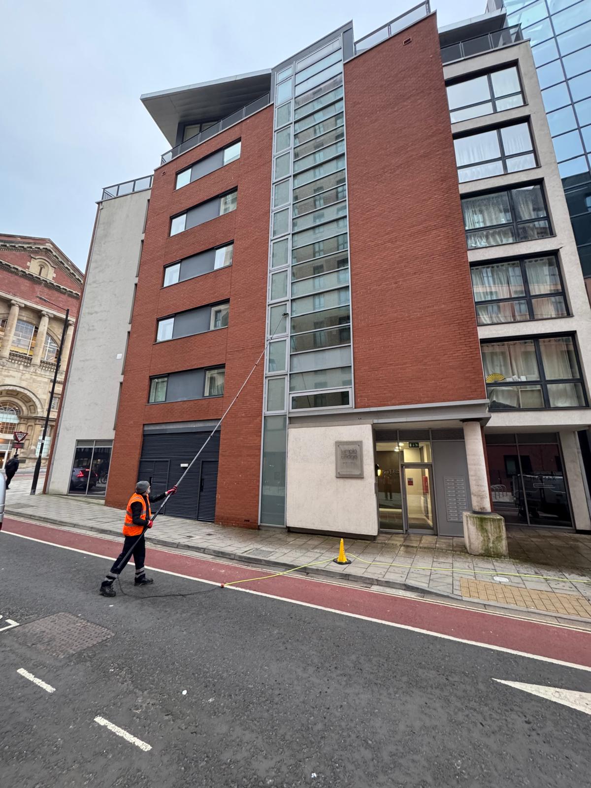 A worker in an orange safety vest uses a long pole for commercial window cleaning on the ground floor of a modern multi-story building with red and gray panels, on a city street.