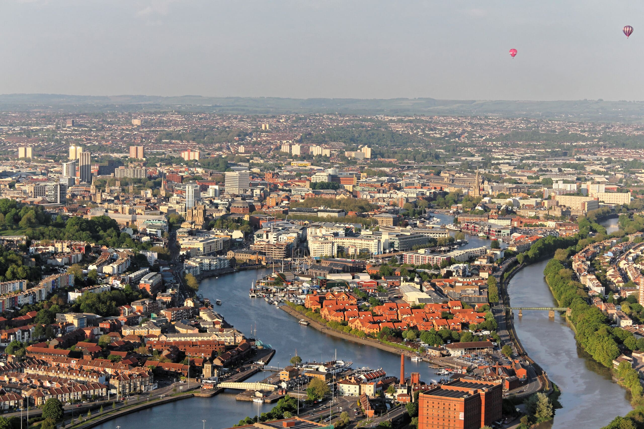 Aerial view of a city with a winding river, clusters of buildings, green areas, and several hot air balloons floating in the sky—perfect for showcasing high level cleaning on tall commercial structures.