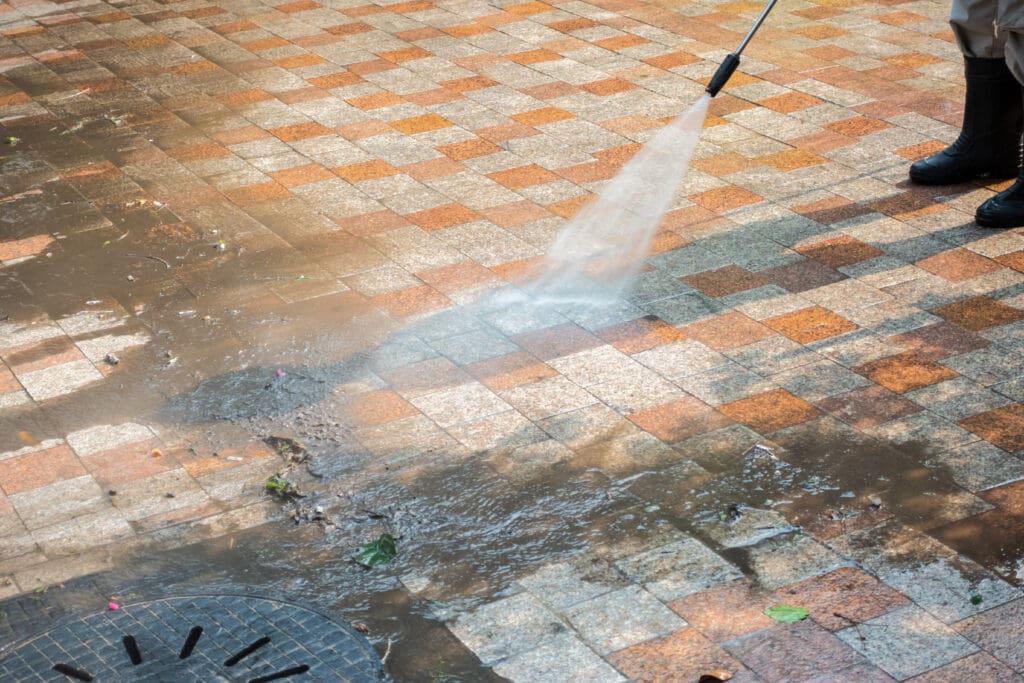 A person wearing rubber boots uses a high-pressure water hose for a thorough driveway jet wash, cleaning a dirty tiled outdoor surface and washing away dirt and debris towards a drain.