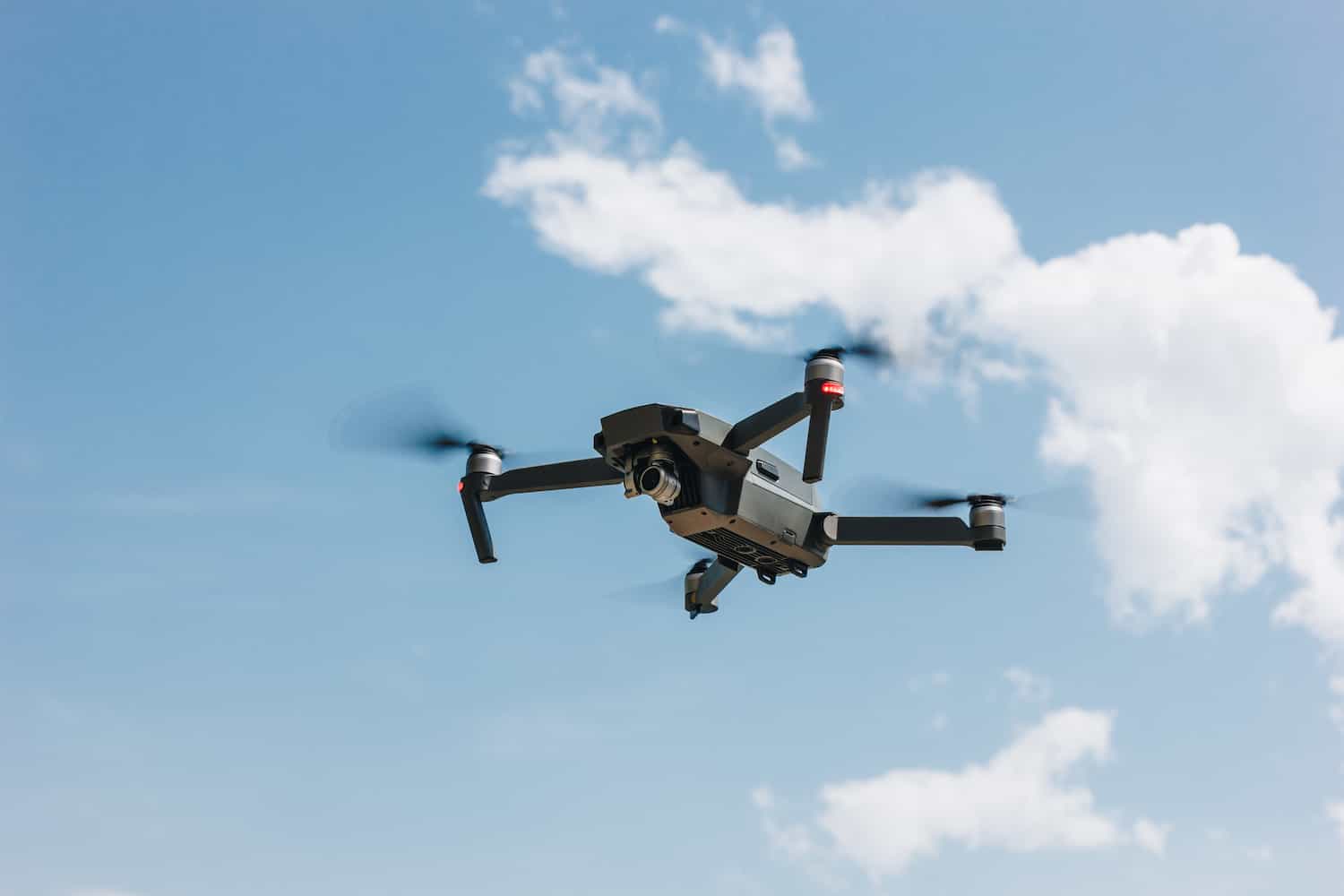 A black quadcopter drone with a camera hovers midair against a blue sky with scattered white clouds.