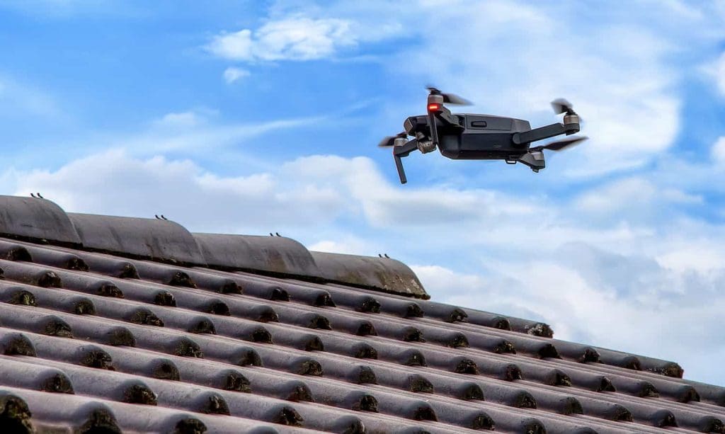 A small black drone with spinning propellers hovers above a tiled roof under a blue sky with scattered clouds.