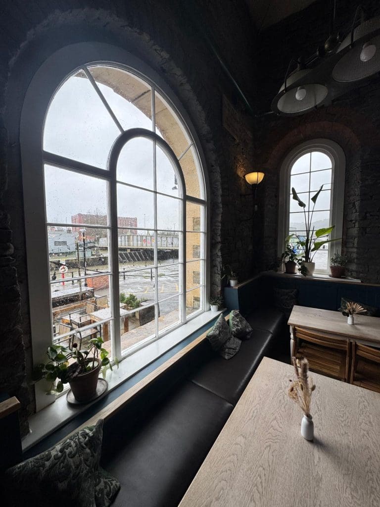 Cozy cafe interior with large arched windows, potted plants, and cushioned seating. Outside, a street and railway tracks are visible on a cloudy day, adding to the relaxed atmosphere.