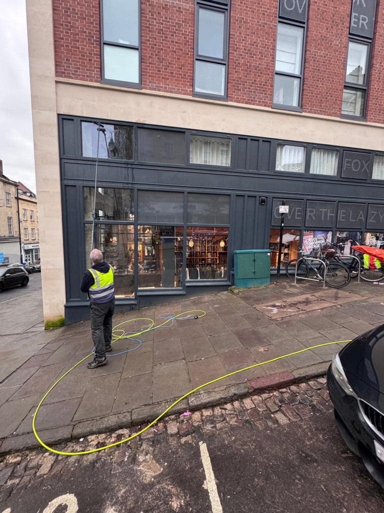 A person wearing a high-visibility vest is cleaning the large windows of a ground-floor restaurant or bar on a city street. A yellow hose is stretched across the pavement, and bicycles are parked nearby.