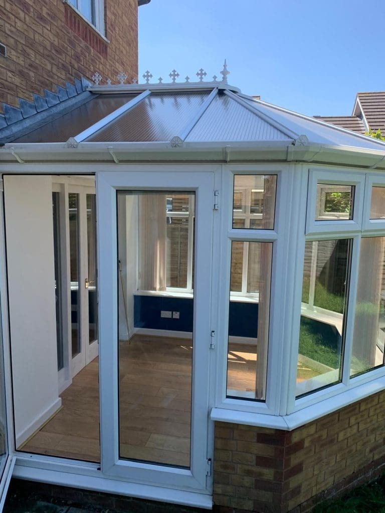A white-framed glass conservatory with a sloped roof is attached to a brick house. The doors and windows are open, revealing a wooden floor and blue walls inside. It is a sunny day with a clear blue sky.