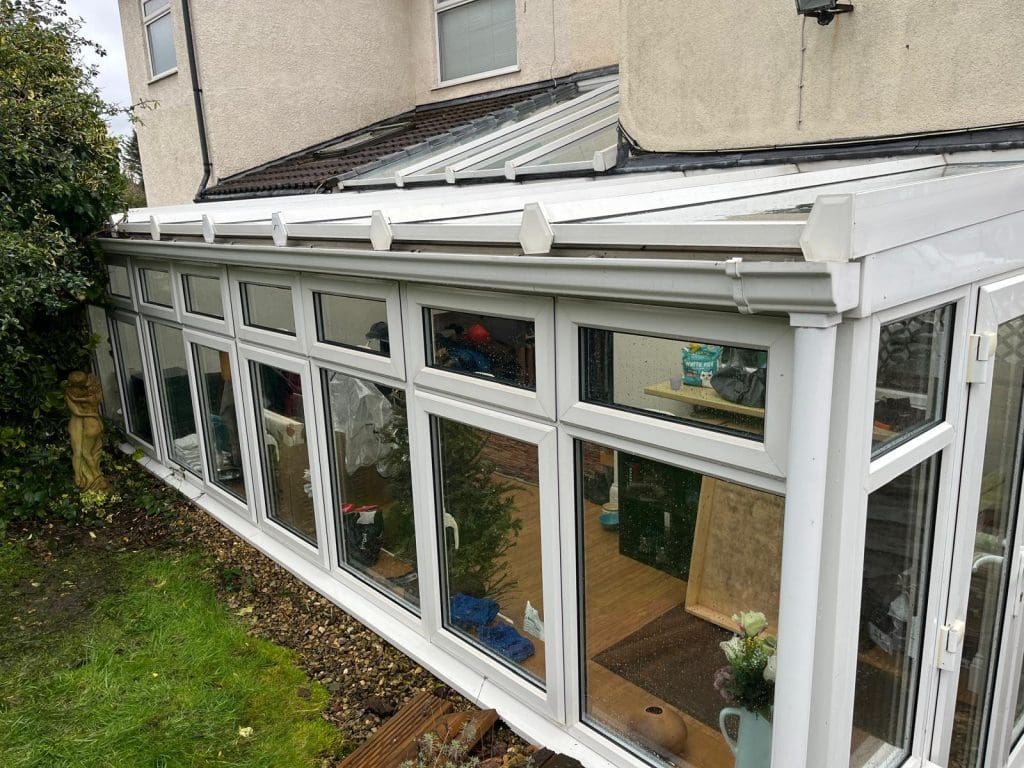 A white conservatory with glass windows and roof panels attached to a house, showing indoor plants and furniture inside. The conservatory is beside a grassy garden with some bushes and a statue.