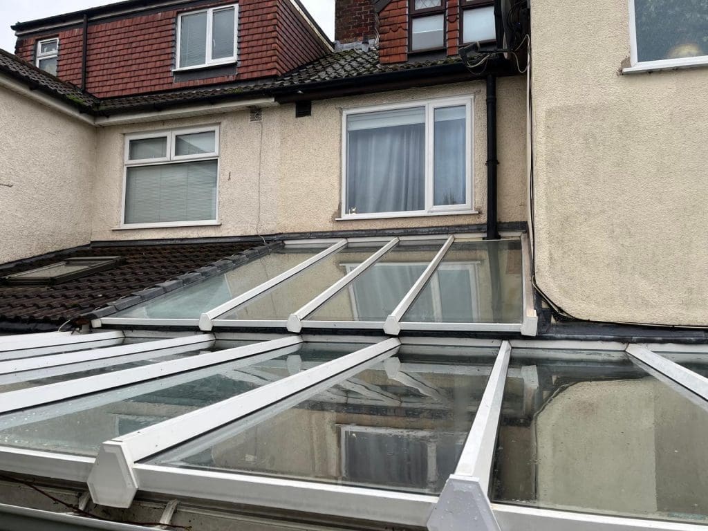 A slanted glass roof extension is attached to the back of a beige house with white-framed windows and a tiled roof. The glass panels reflect the overcast sky.