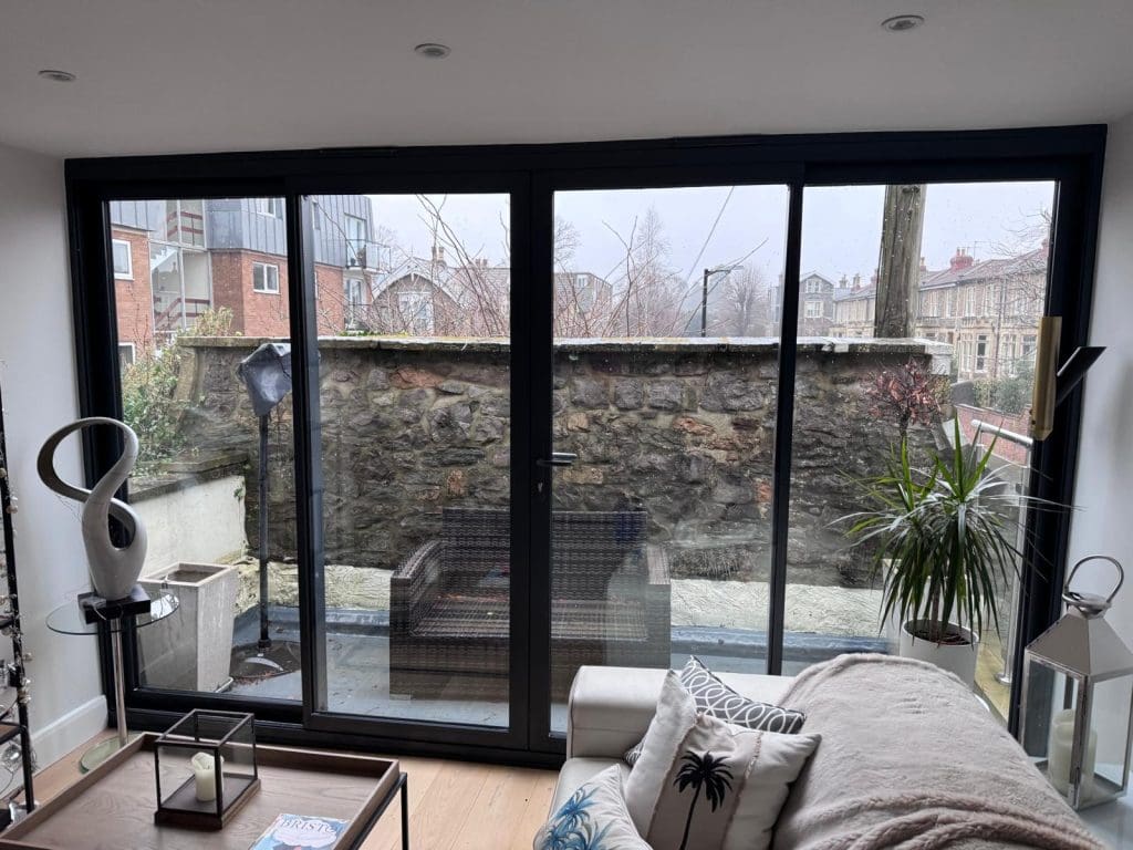 A modern living room with glass doors opening to a small patio with wicker furniture, a tall plant, and a stone wall. Buildings and leafless trees are visible in the background on a cloudy day.