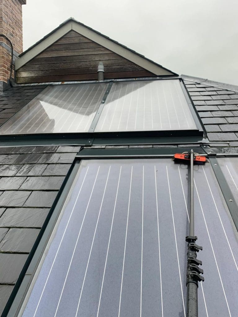 A close-up view of solar panels installed on a sloped roof, with a long-handled cleaning tool being used to wash the panels on a cloudy day.