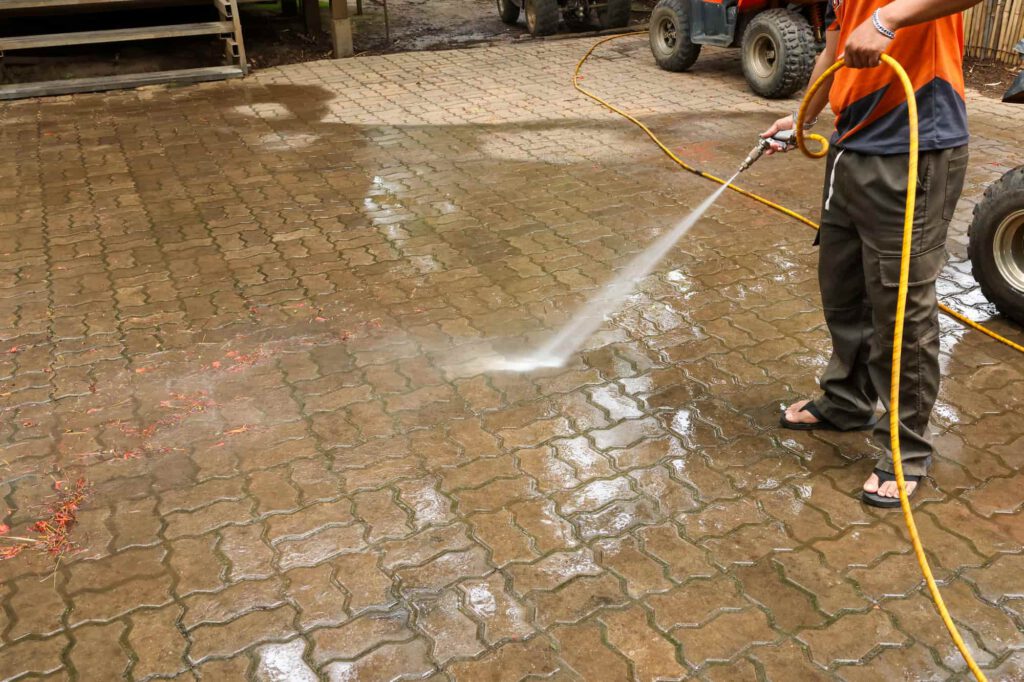 A person in sandals uses a pressure washer to clean a brick-paved outdoor surface, spraying away dirt and debris.