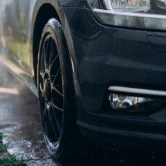 Close-up of a black car’s front right tire and headlight, partially wet, with water on the ground and grass visible at the left edge.