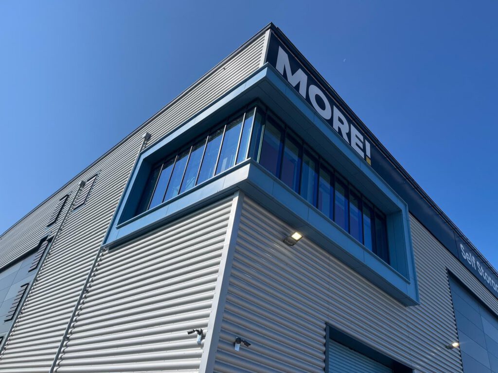 A modern industrial building with light gray metal siding, a blue-framed window, and “MORE!” written in bold white letters on the upper corner, seen against a clear blue sky.