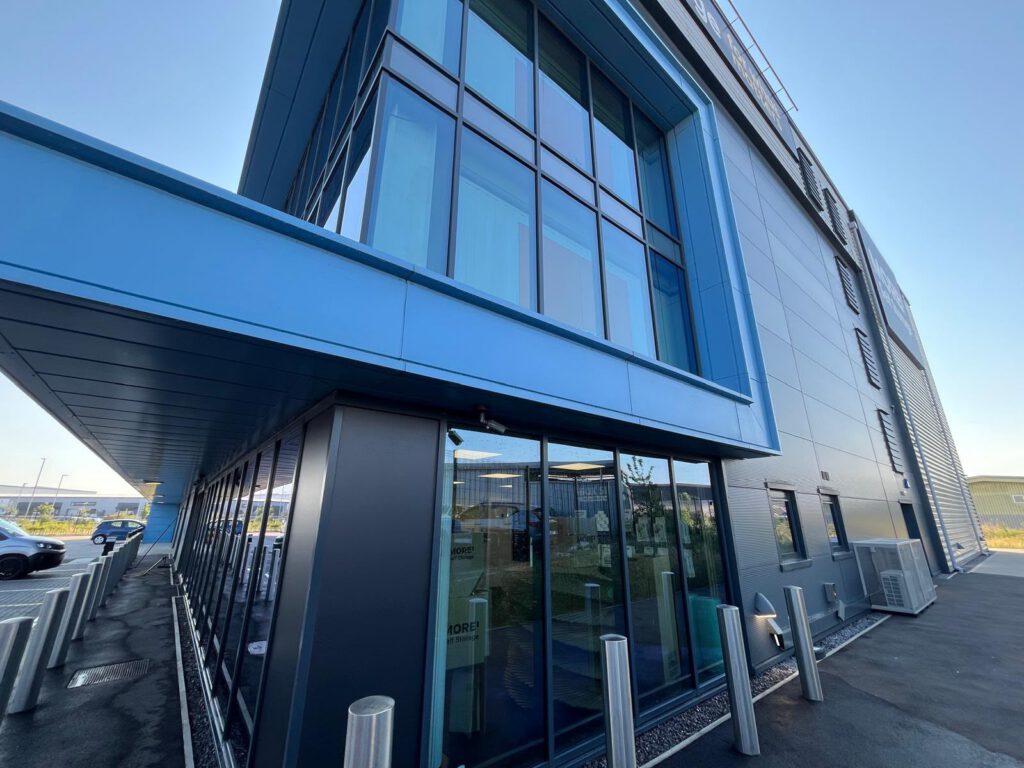 Modern glass and metal building with large windows, blue trim, and reflective surfaces. Metal bollards line the sidewalk, and part of the parking lot is visible on a clear, sunny day.