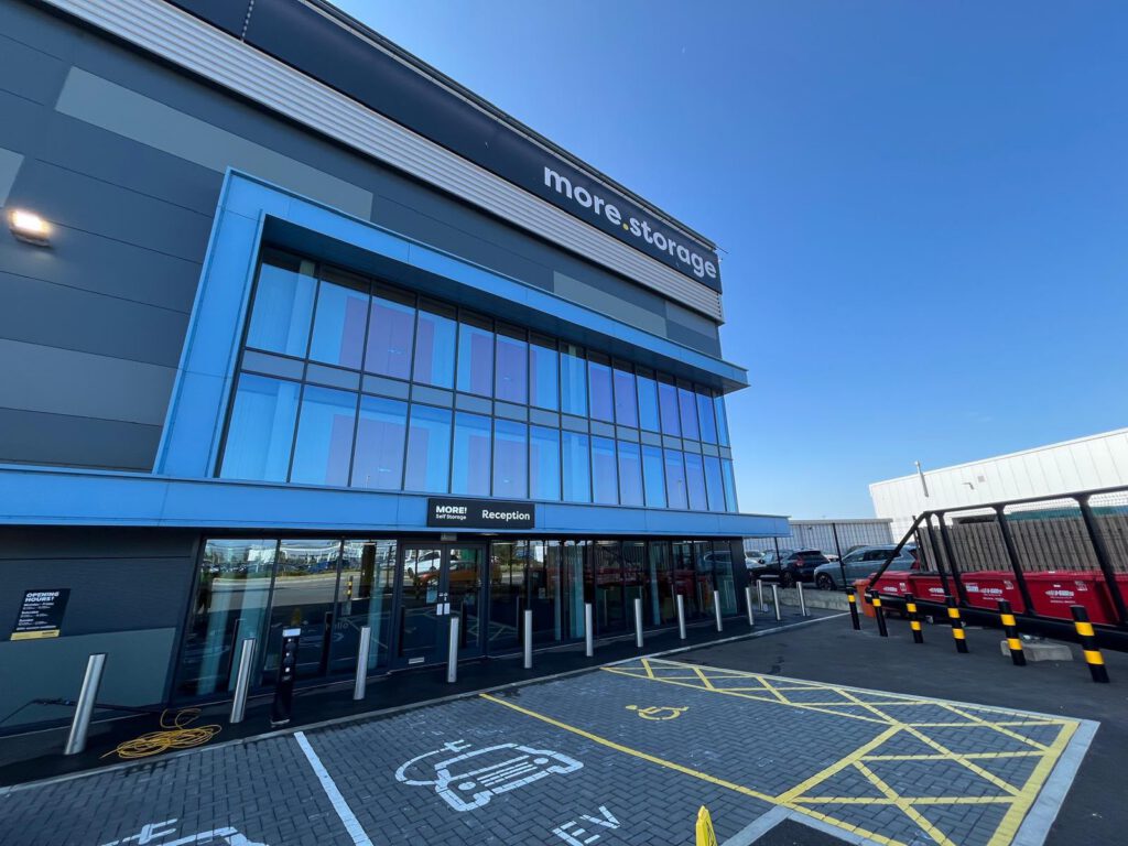 A modern self-storage building with large glass windows, labeled "more.stoorage." The entrance says "Reception." There are parking spaces and a few red vehicles parked outside on a sunny day.