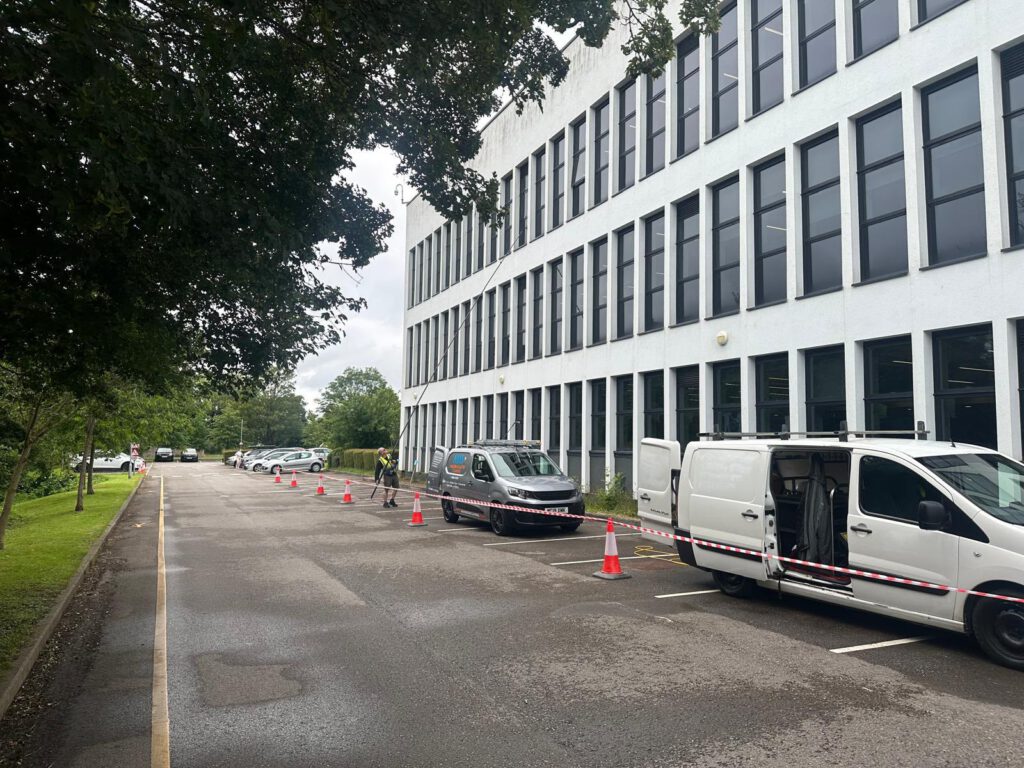 A row of parked white vans beside a modern, multi-story office building, with orange cones marking off several parking spaces on a cloudy day.