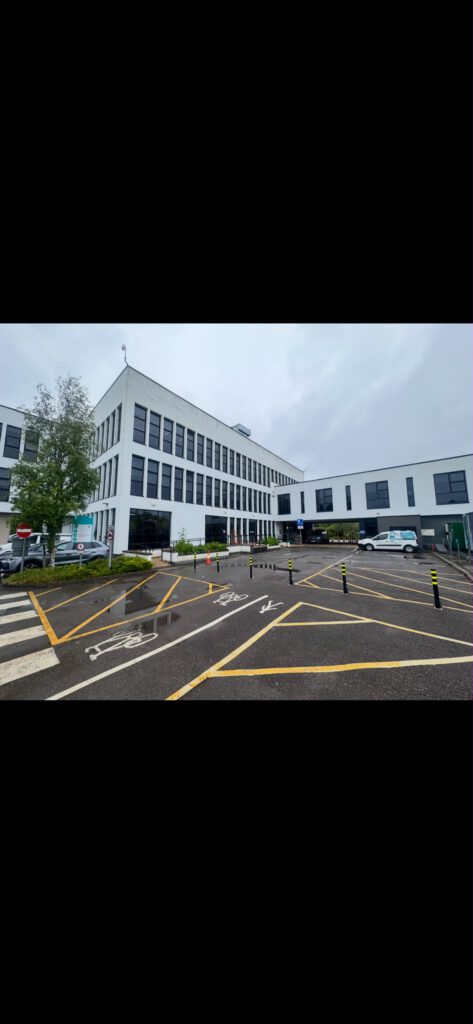 A modern white office building with many windows, a parking lot with marked spaces and bollards in the foreground, and a cloudy sky above.