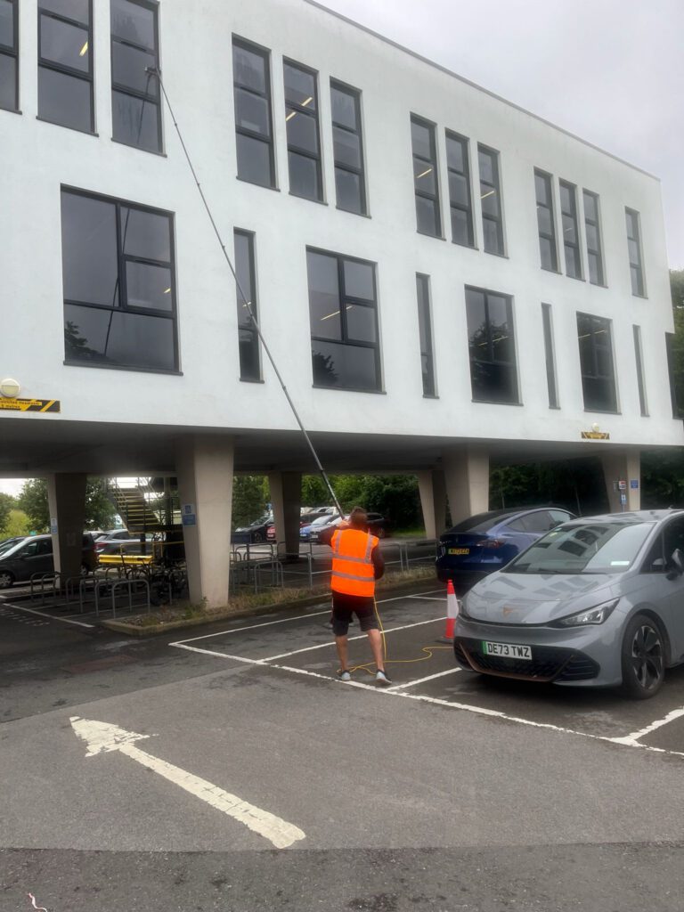 A person in an orange vest uses a long pole to clean the upper windows of a white building in a parking lot with cars and bike racks visible.
