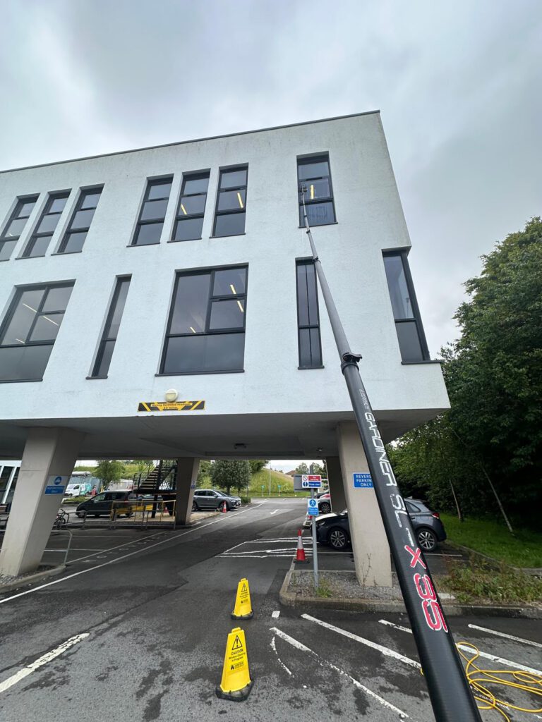 A long pole with a brush is being used to clean third-floor windows of a white building. Yellow caution signs are placed on the wet ground below, near parked cars.