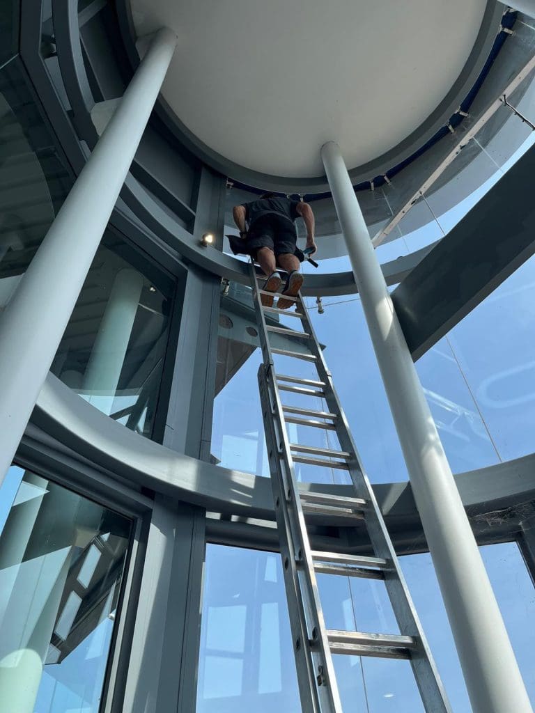 A person standing near the top of a tall metal ladder inside a modern glass building, working close to the ceiling with sunlight streaming through large windows.